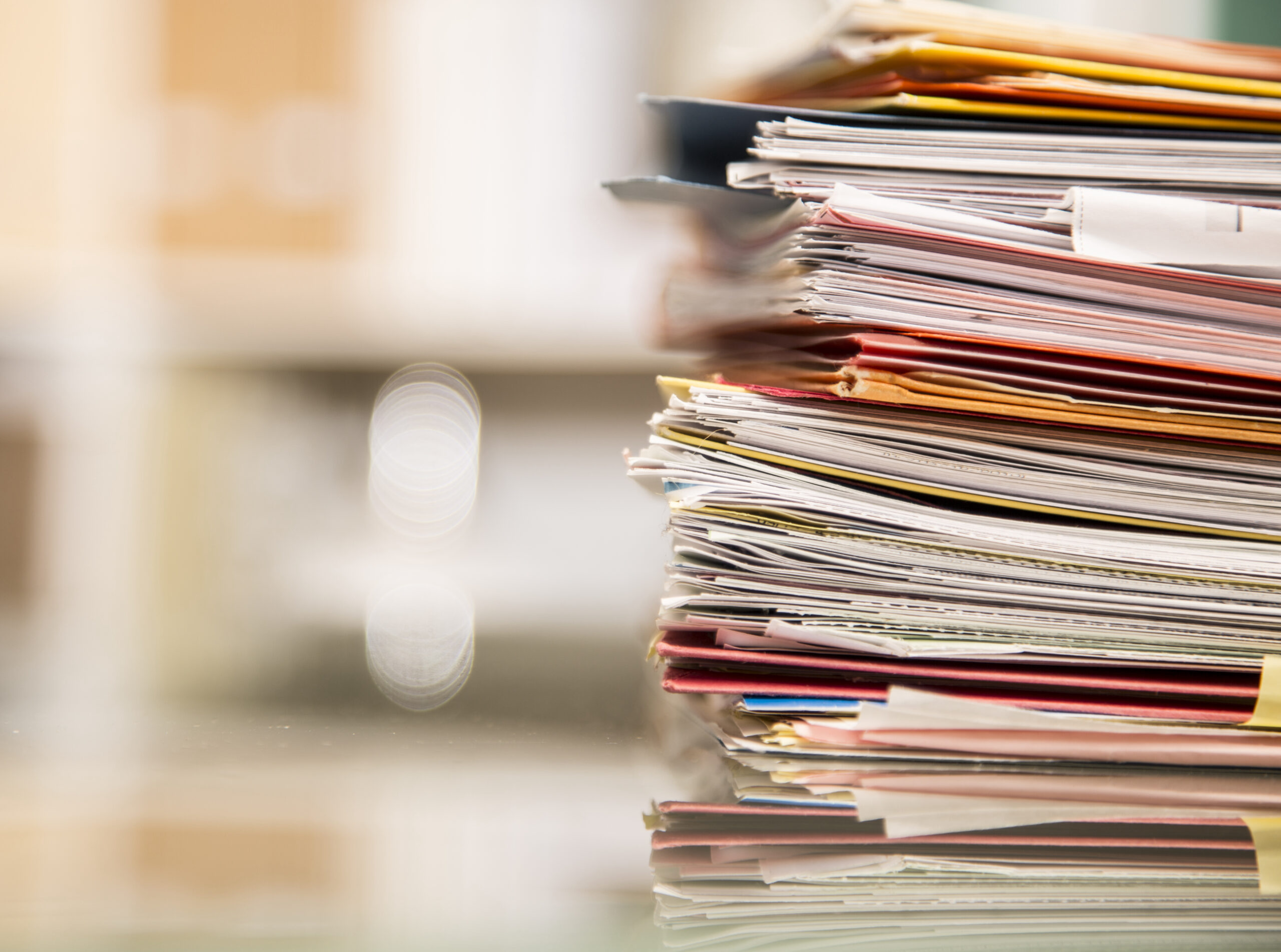 Large stack of file folders, documents, paperwork piled on glass top desk in office.  Bookshelves in background.